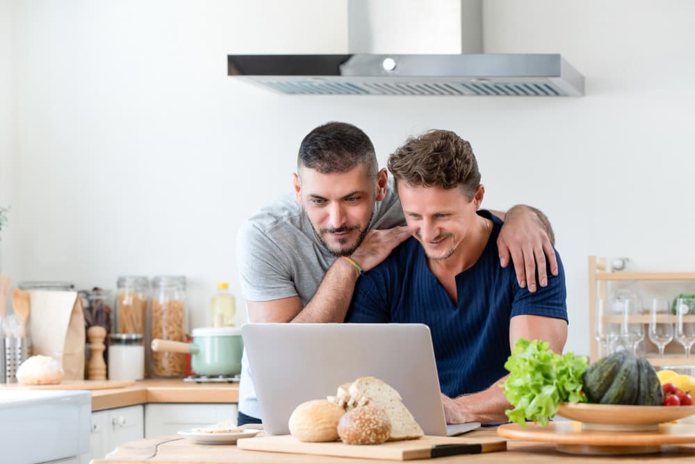 Two men in a kitchen smiling at a laptop together