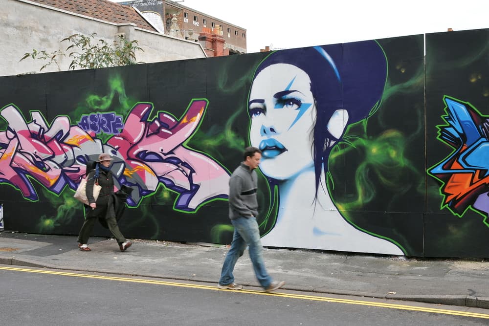 UK man and woman walk by graffiti mural on street, Bristol