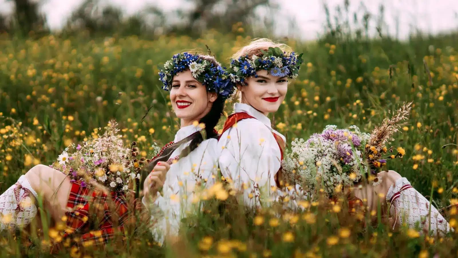 Two women in Latvian folk dress and flower crowns sitting in a summer meadow