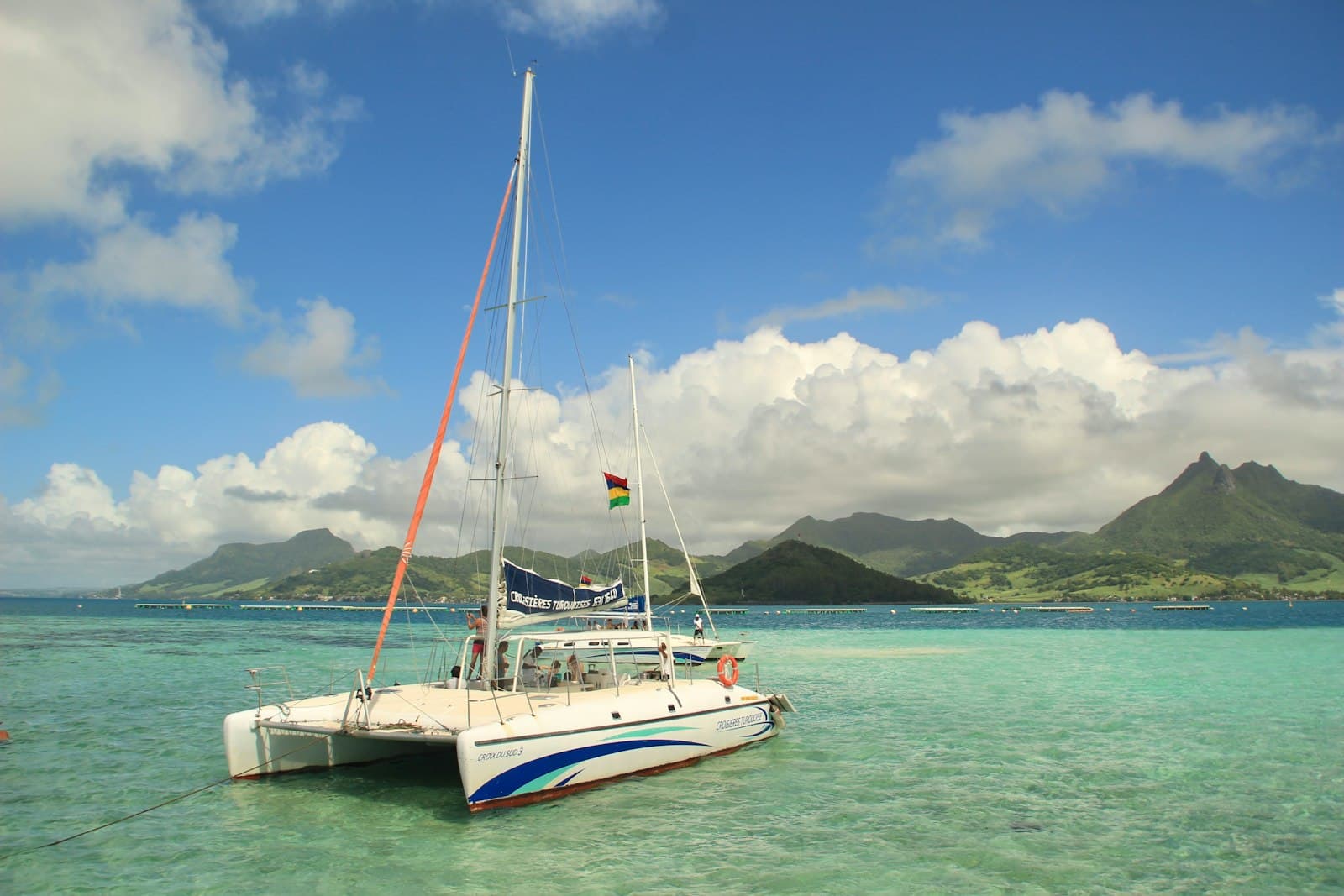 A catamaran flying the Mauritian flag anchored in a turquoise lagoon with green mountains behind
