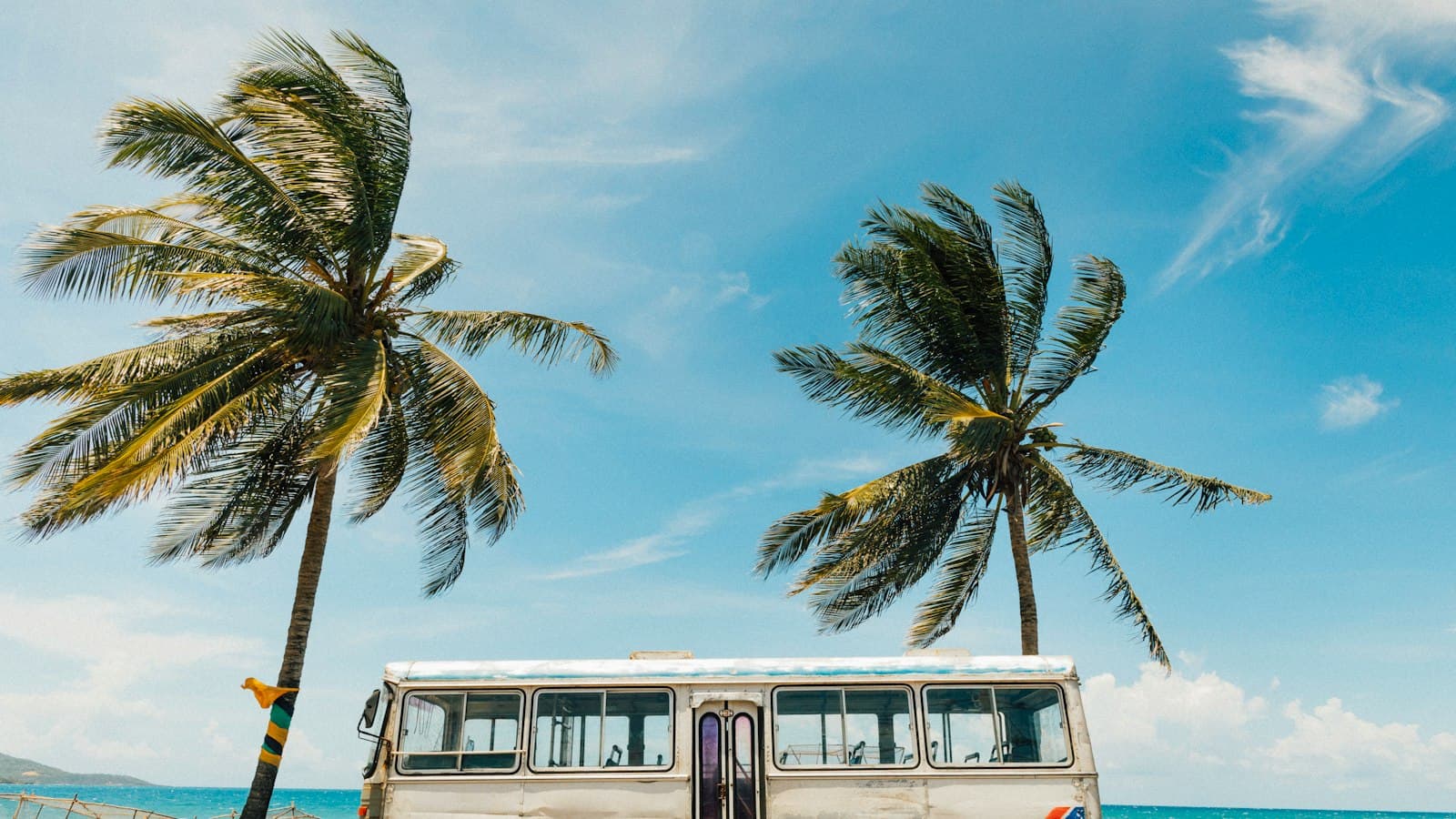 An old white bus parked by the sea under two palm trees, a Jamaican flag stripe on the pole