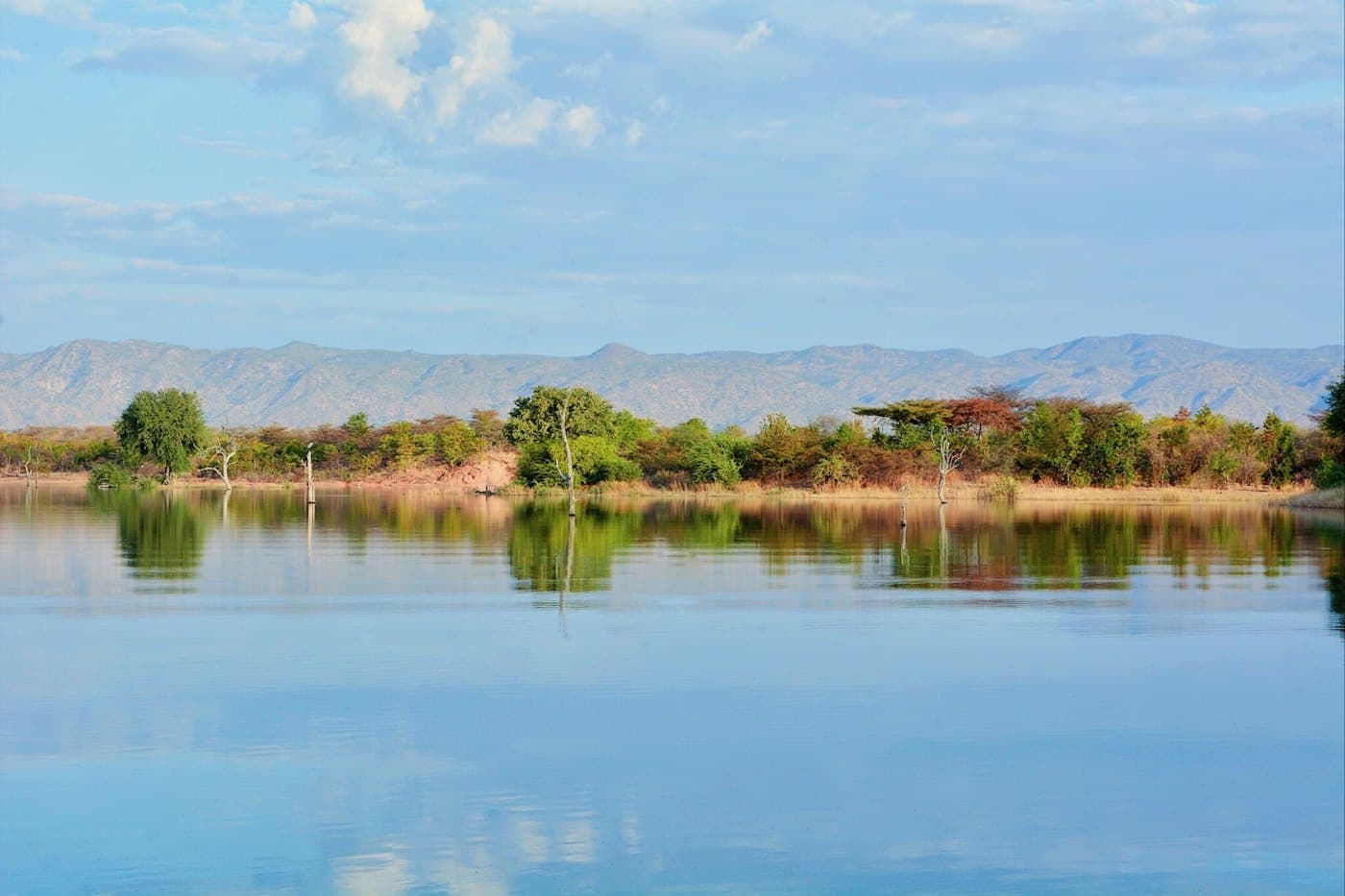 A calm lake reflecting green trees, with dry hills behind under a pale blue sky