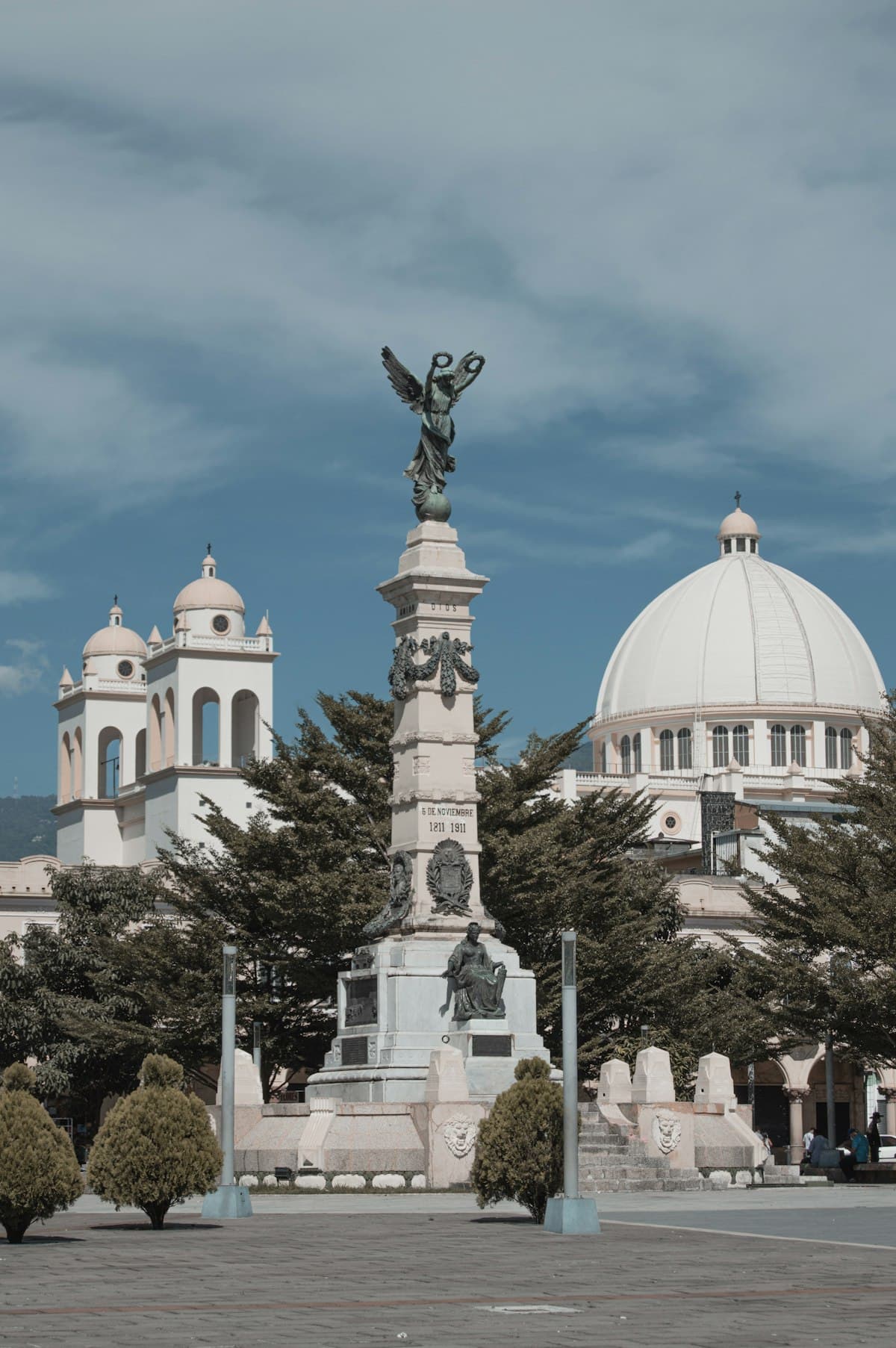Liberty monument in front of the white dome and bell towers of a church on a plaza in downtown San Salvador