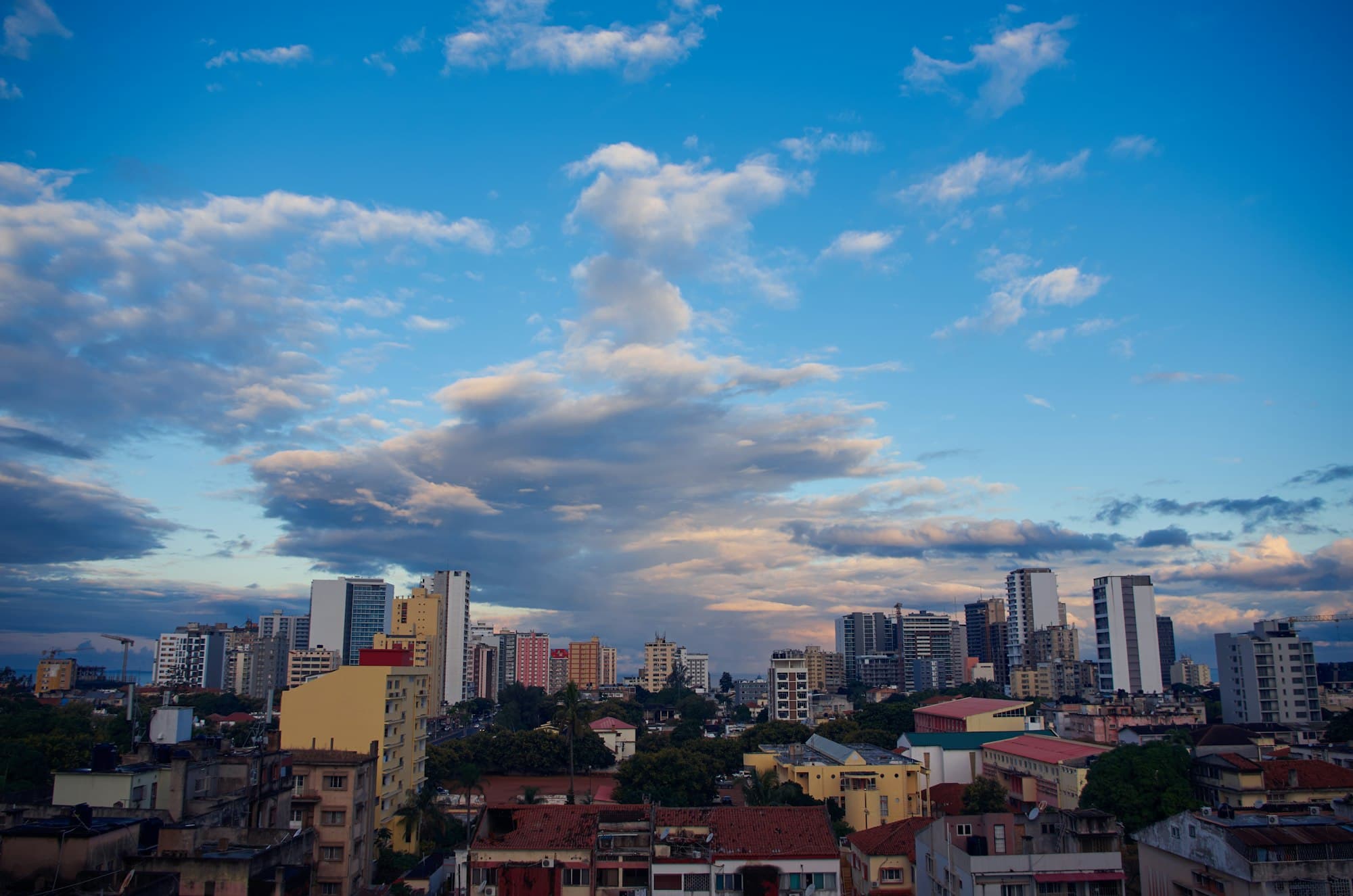 Maputo's skyline of apartment towers and red-roofed low-rise buildings under a blue evening sky