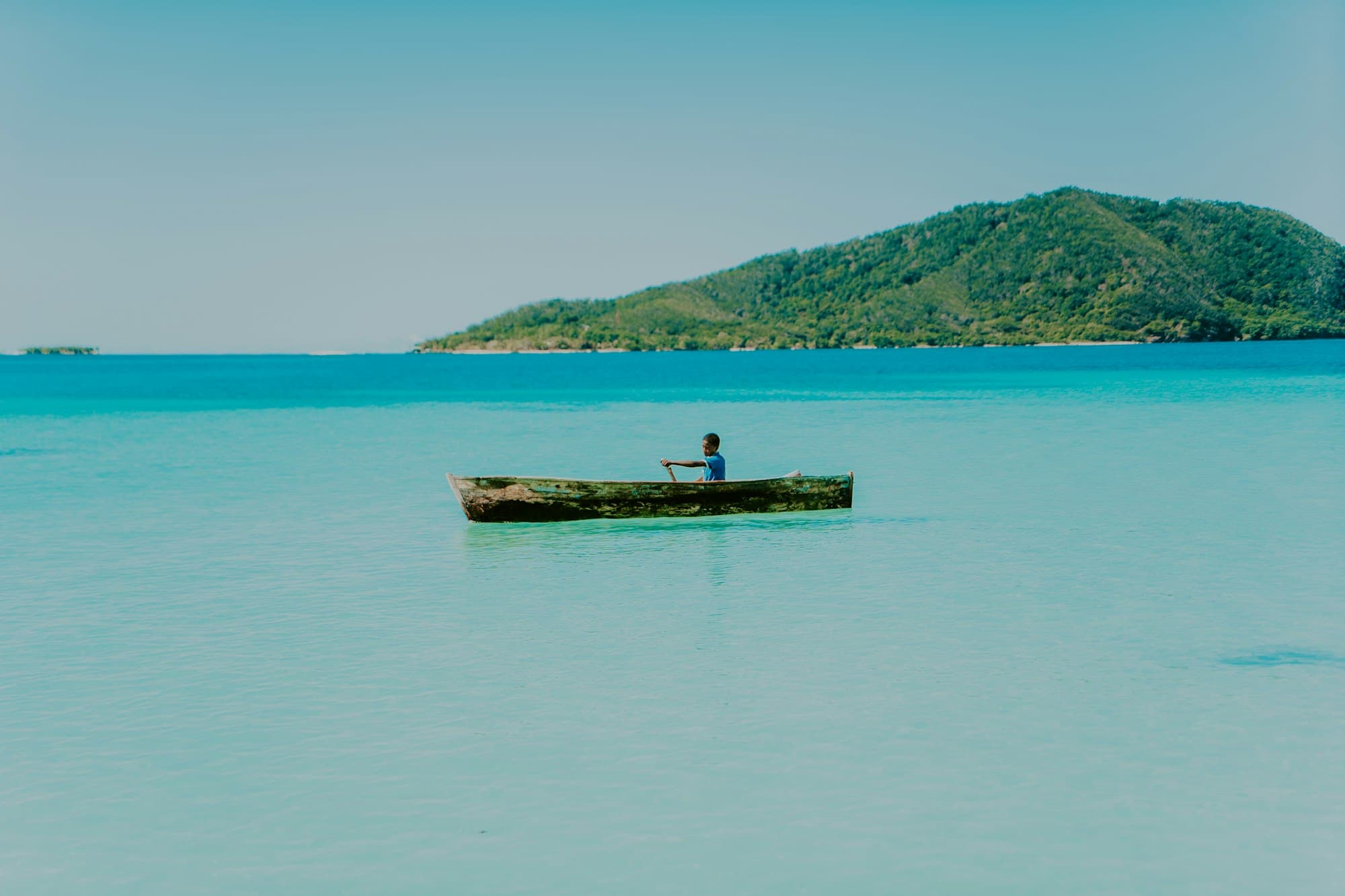 A boy paddling a wooden canoe on turquoise water off a green island in Honduras