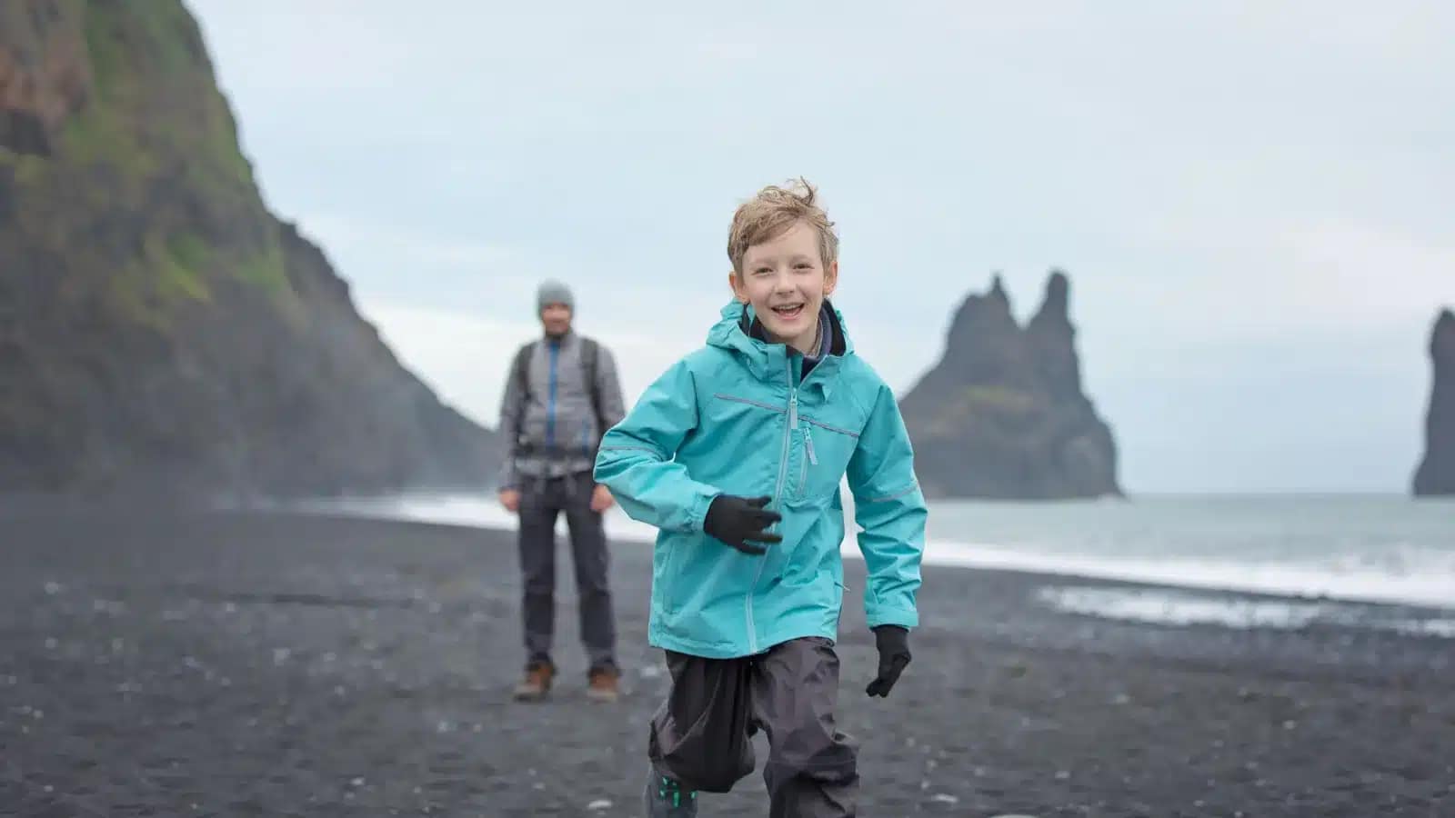 A boy in a blue jacket runs along a black sand beach in Iceland, sea stacks behind him