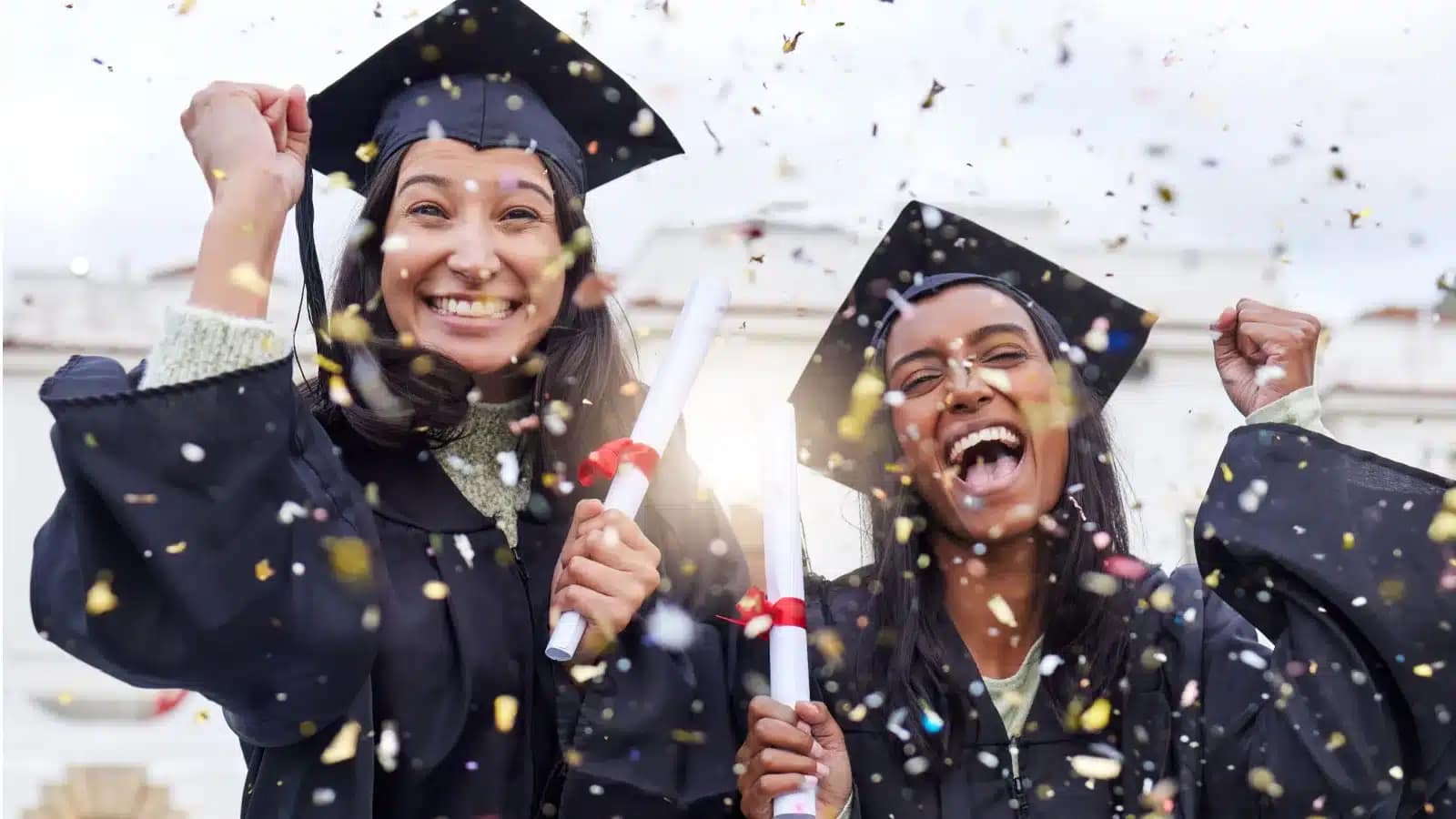 Two graduates in caps and gowns celebrating under falling confetti