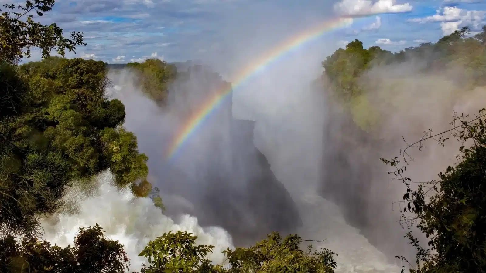 A rainbow arcs over the spray of Victoria Falls between forested cliffs
