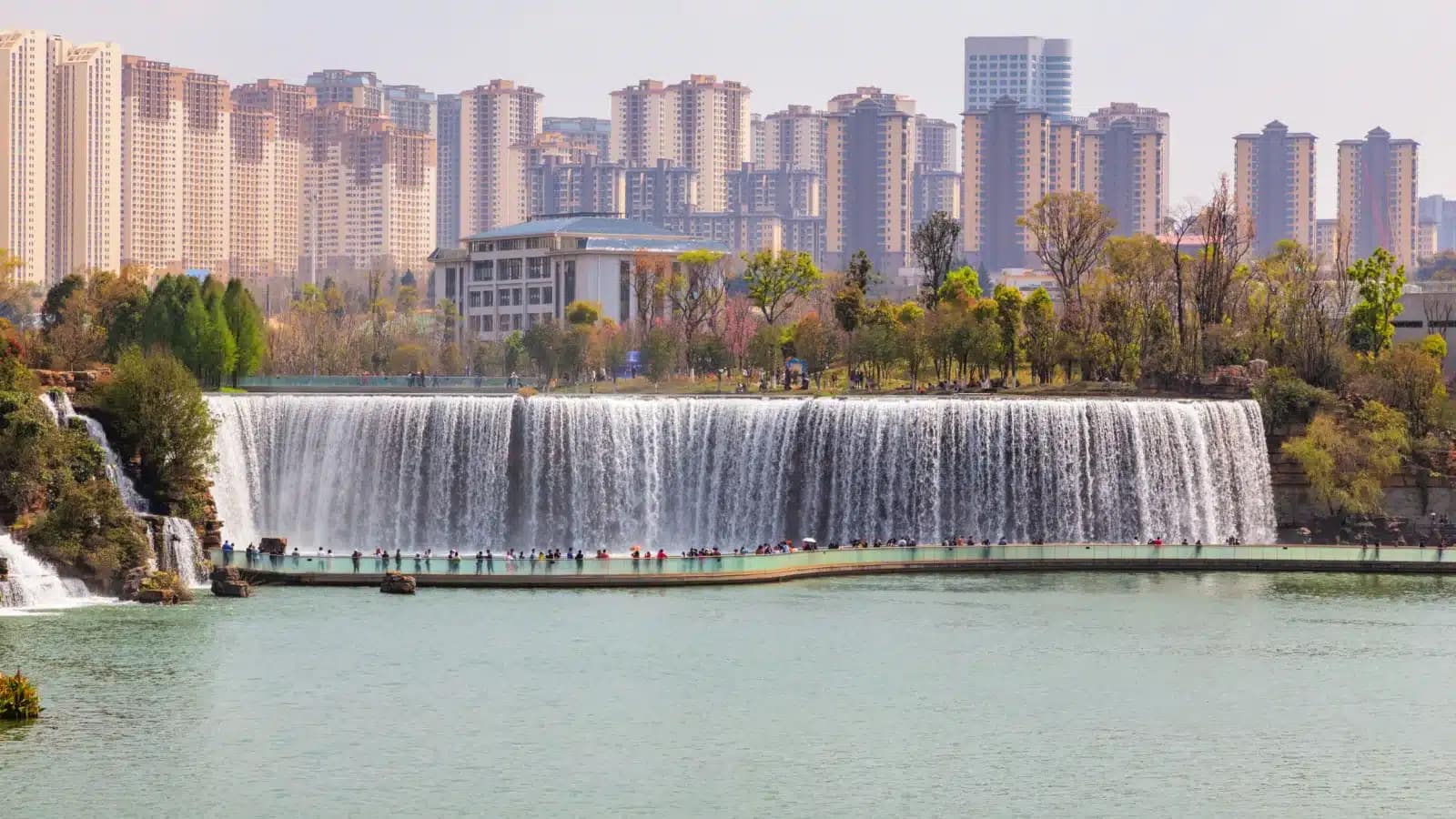 A wide artificial waterfall in a Kunming park, with apartment towers behind it