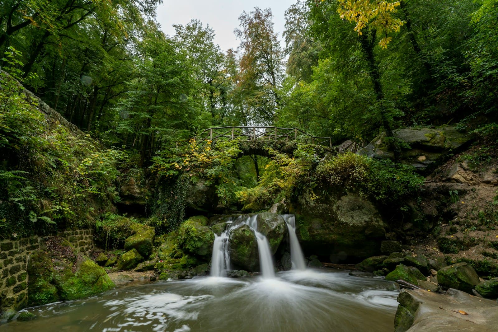 A small waterfall under a rustic wooden footbridge in a Luxembourg forest
