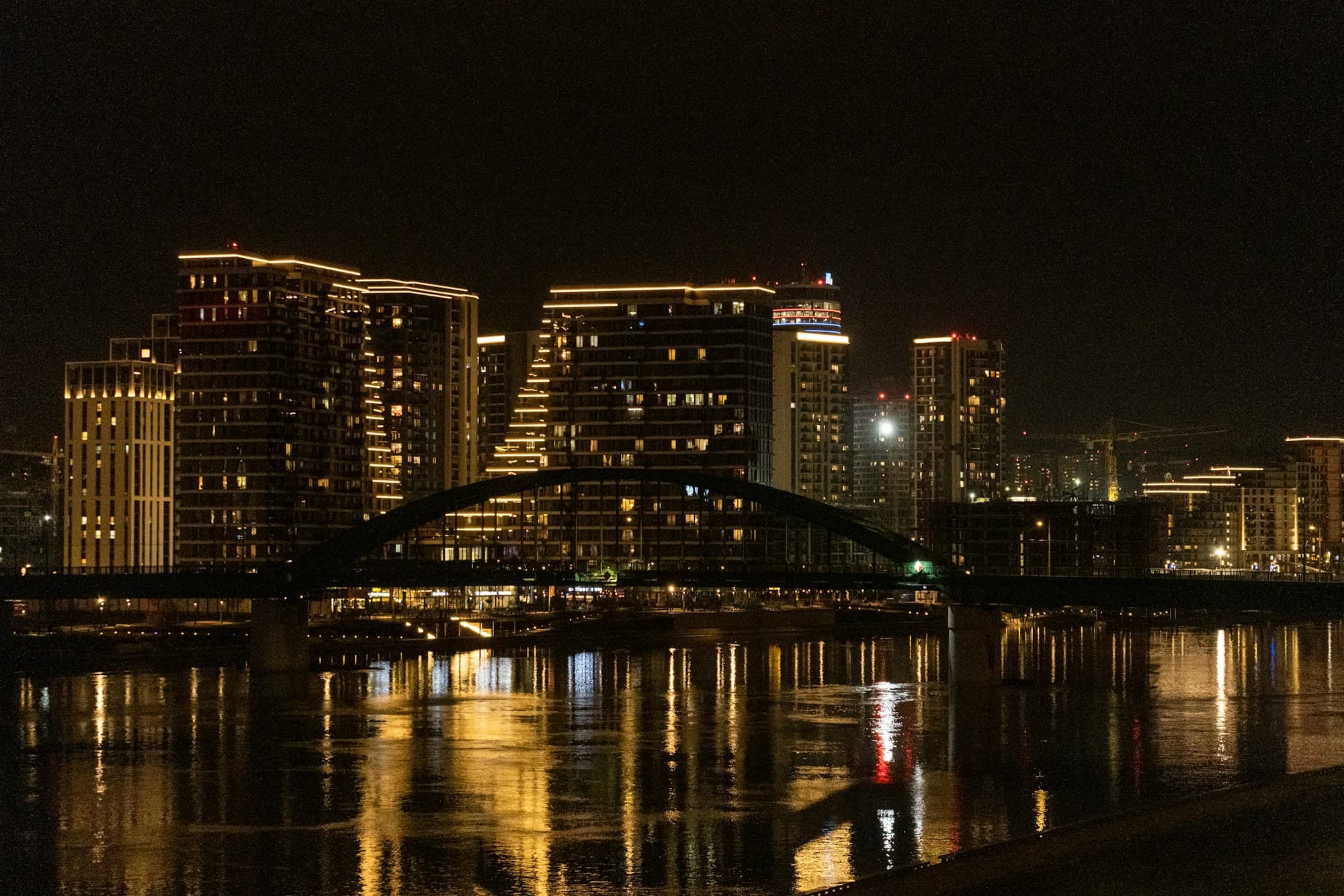 Belgrade's new riverside towers lit up at night, reflected in the Sava beside an arched bridge
