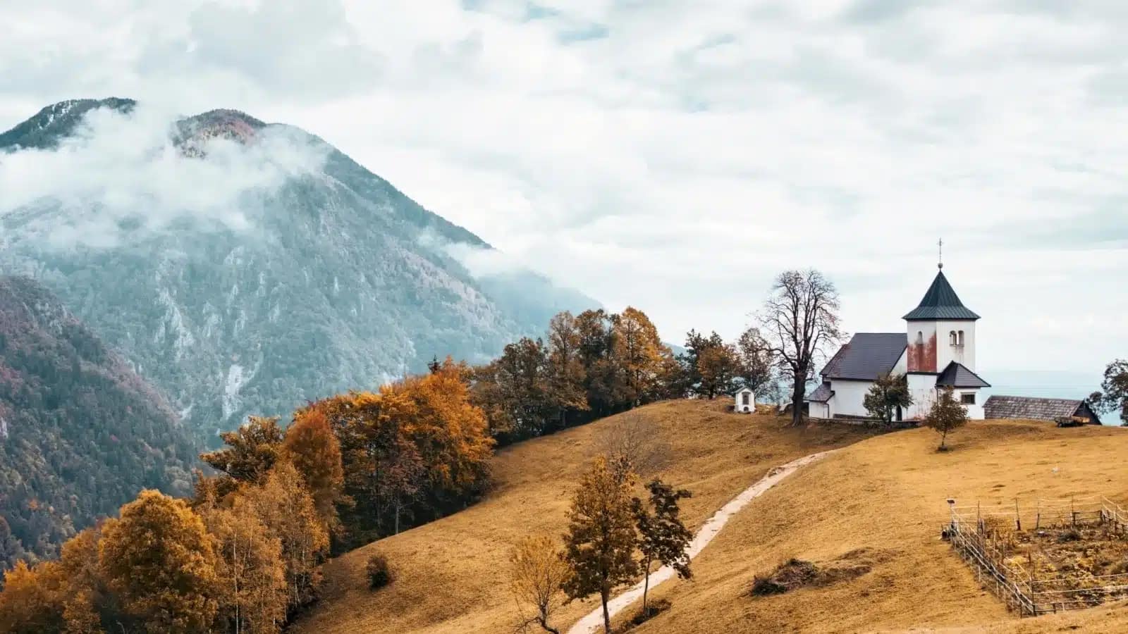 A small white church on an autumn hillside below mist-covered Alpine peaks in Slovenia