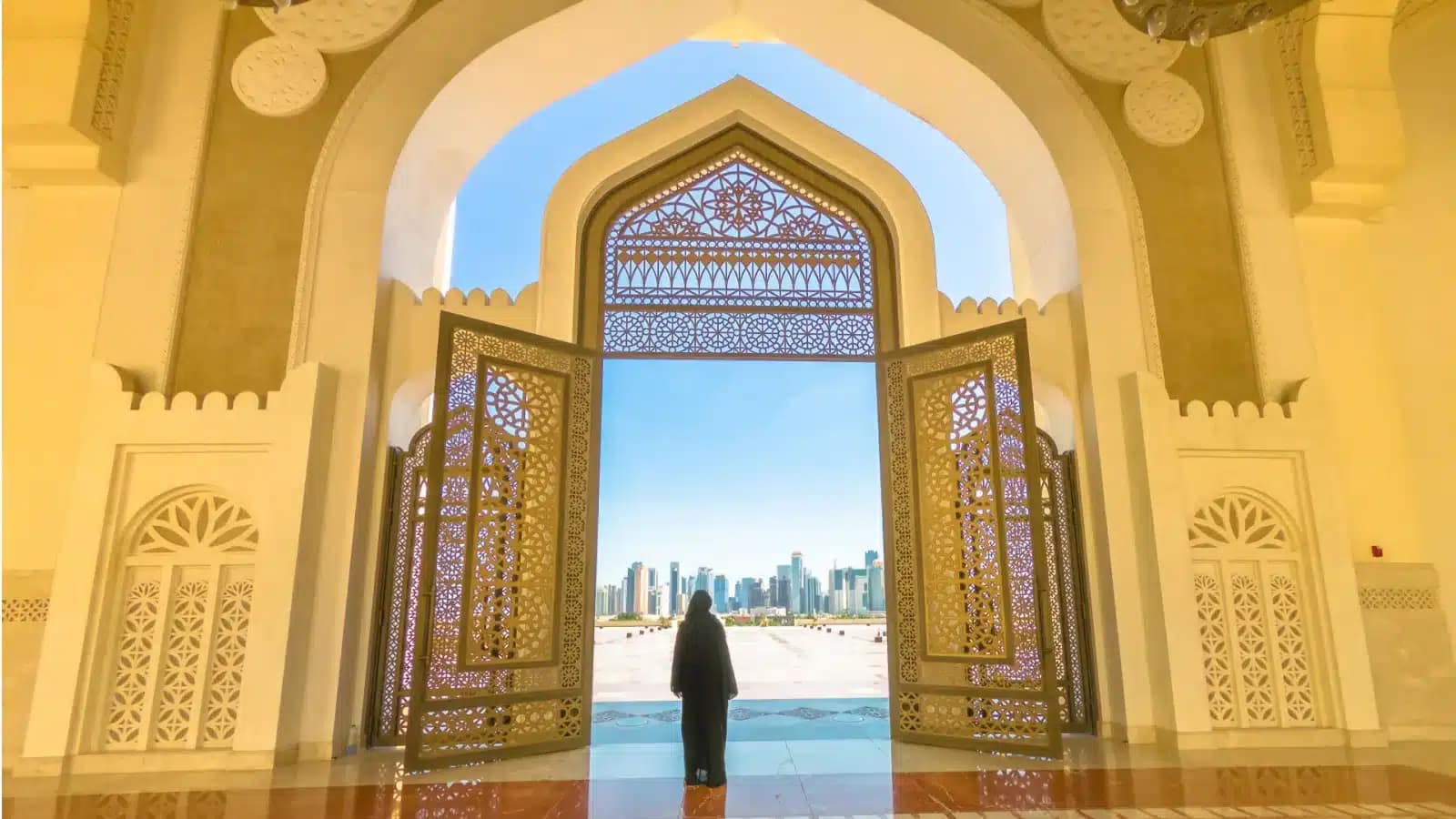 A woman looks through the carved doors of a Doha mosque toward the West Bay skyline
