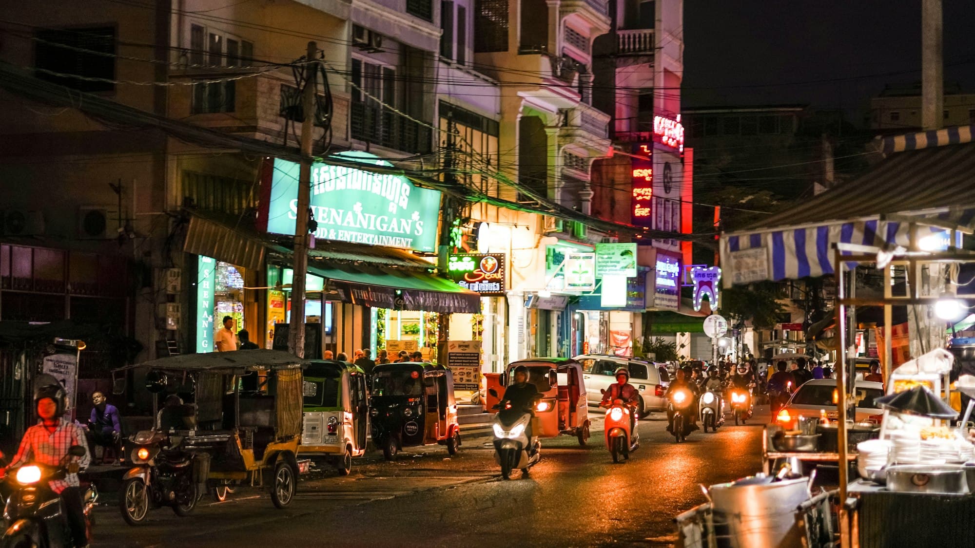 Motorbikes and tuk-tuks on a neon-lit Phnom Penh street at night