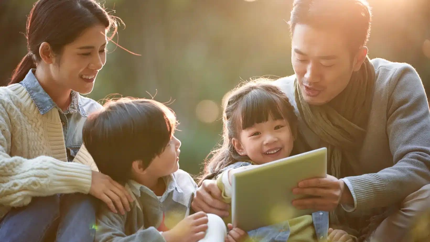 A family of four sits outdoors in soft evening light looking at a tablet together