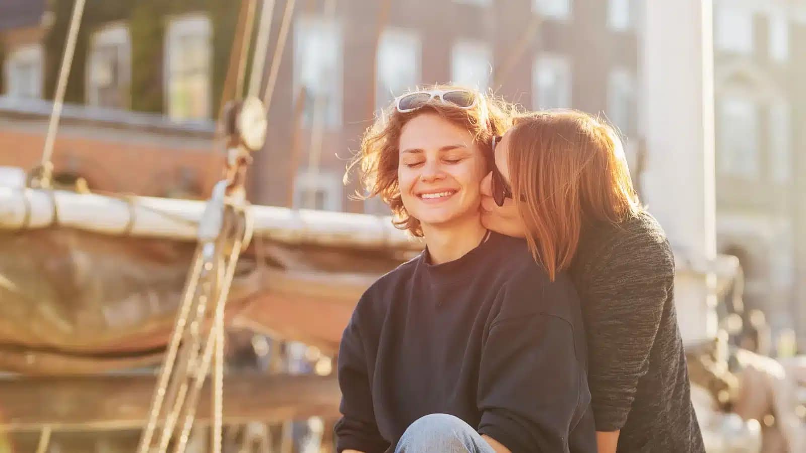 Two women smiling together beside a moored sailboat on a Copenhagen waterfront