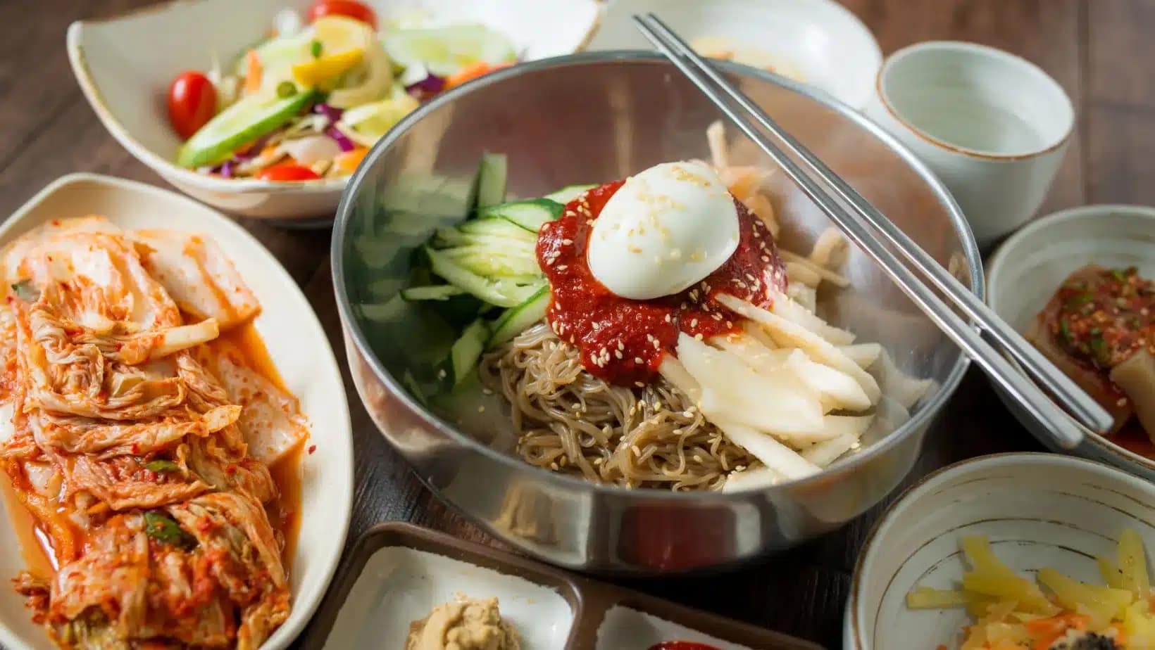 Cold spicy noodles topped with egg surrounded by kimchi and Korean side dishes