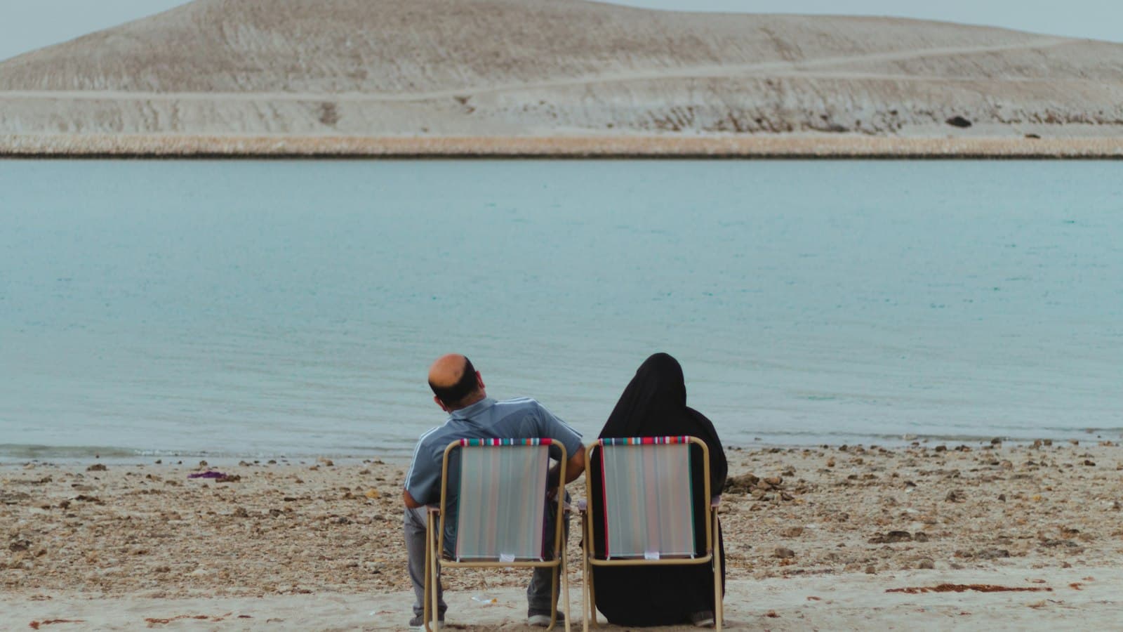 An older couple in beach chairs looking across calm water to a sandy island in Bahrain