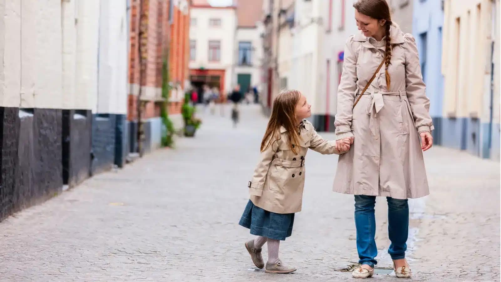 A mother and young daughter walking hand in hand down a quiet Belgian street