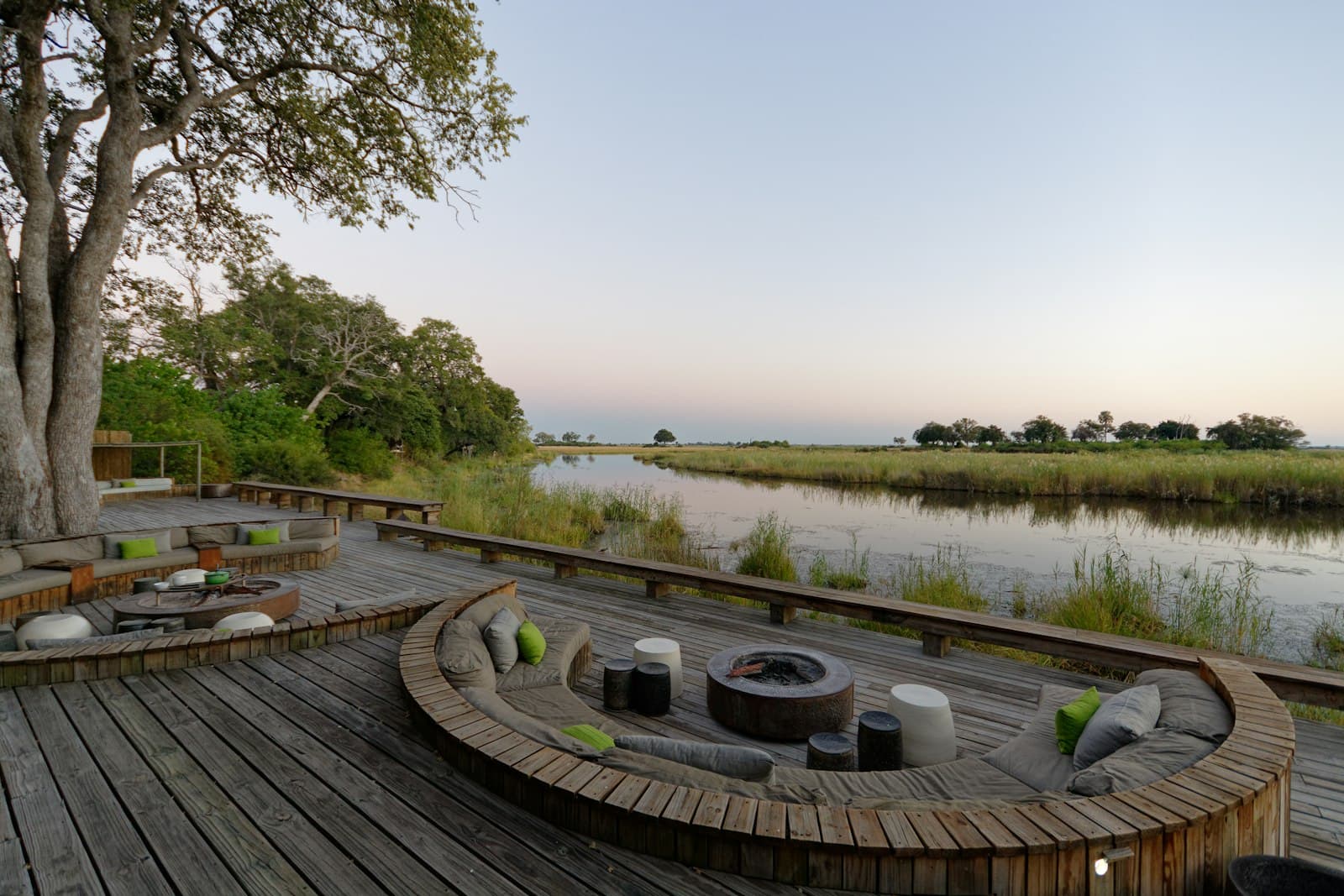A wooden lodge deck with a sunken fire pit beside a quiet Okavango channel at dusk