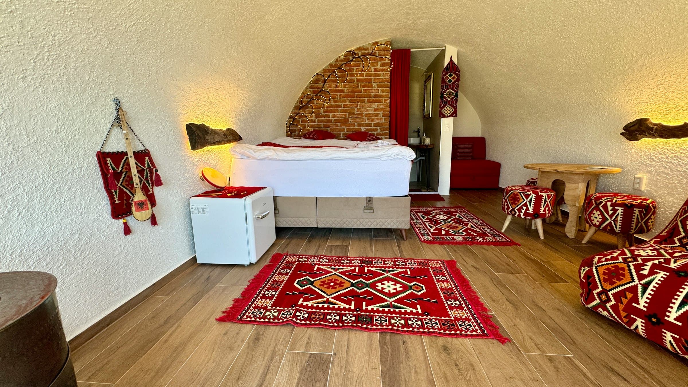 Guest room with red Albanian-pattern rugs, stools, and an embroidered wall hanging