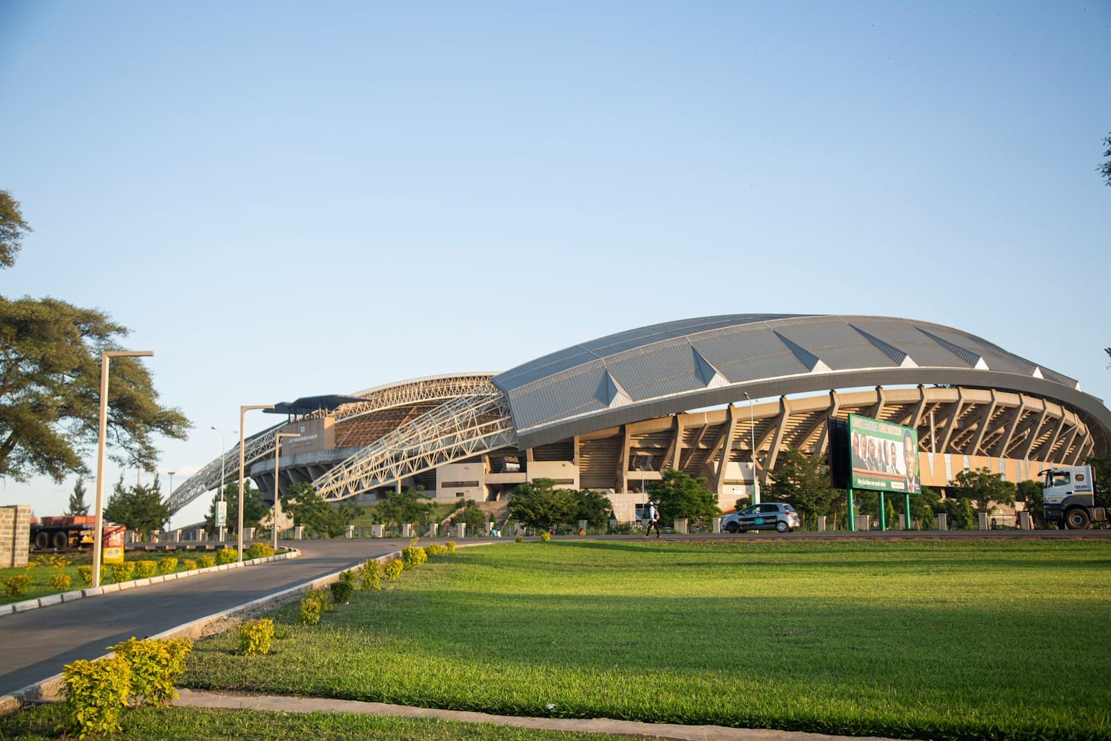 A large modern stadium with a curved roof behind a green lawn in Zambia