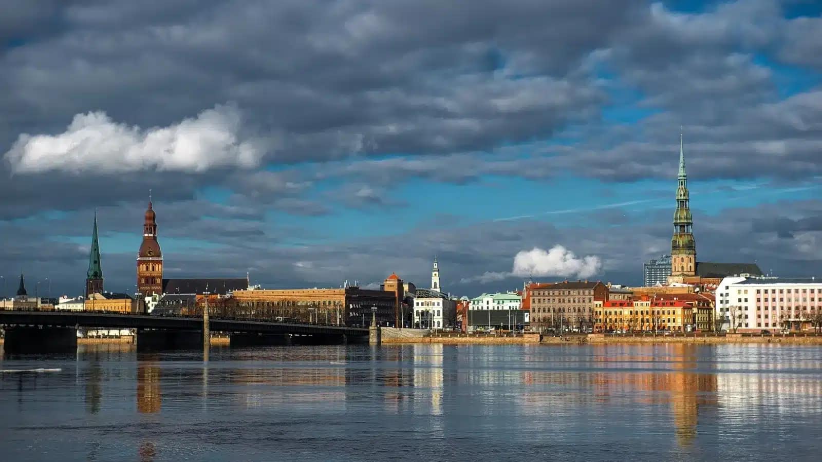 Riga's church spires and riverfront reflected in the Daugava River under a cloudy sky