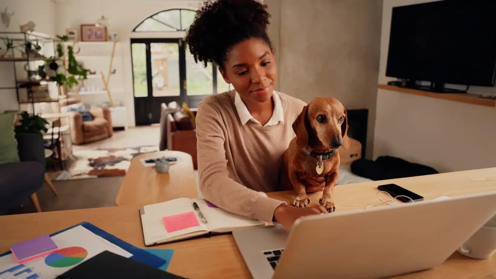 A self-employed woman at her home desk with a laptop, notes and her dog
