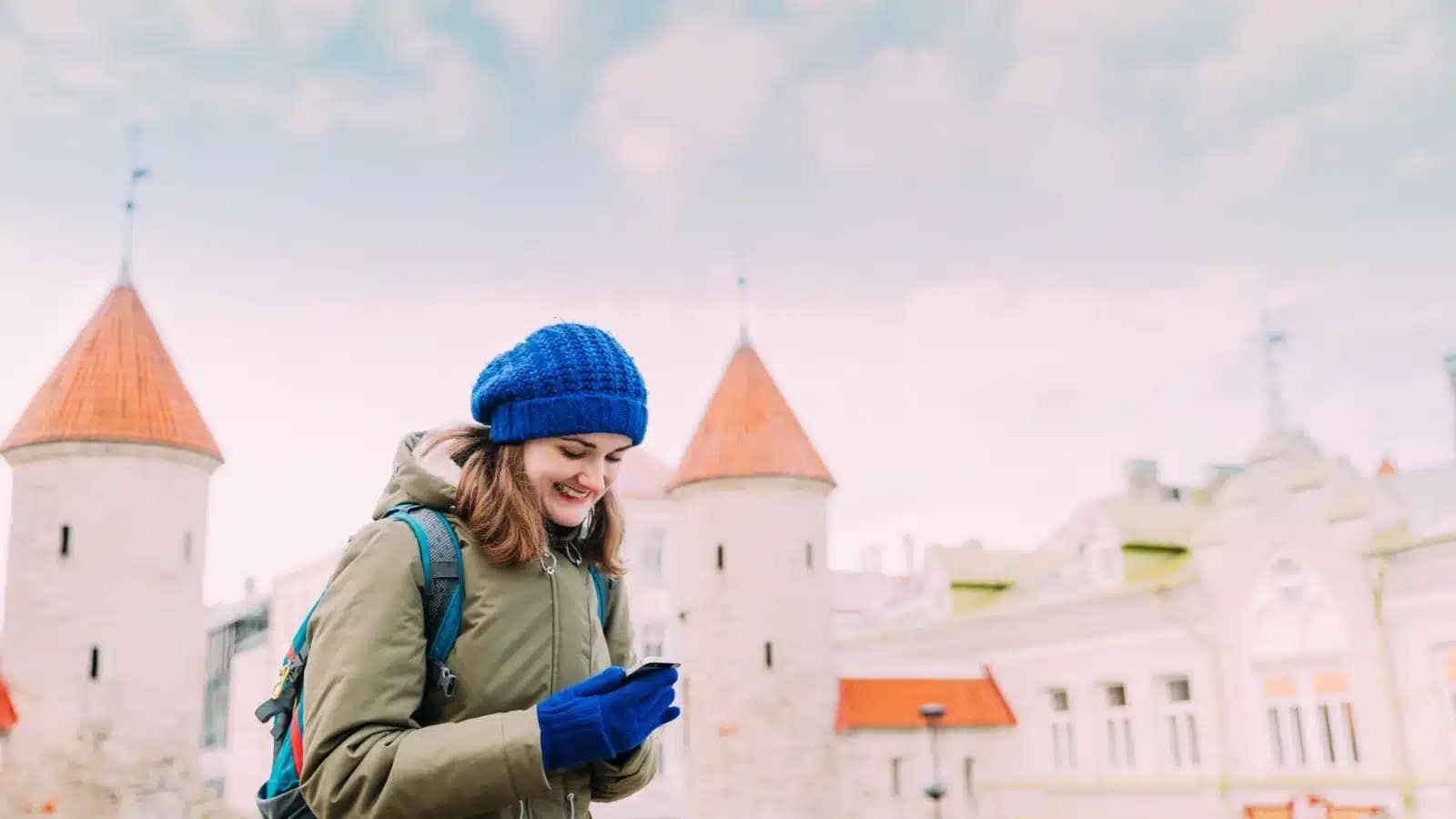 A woman in a knit hat checks her phone in front of Tallinn's Viru Gate towers in winter