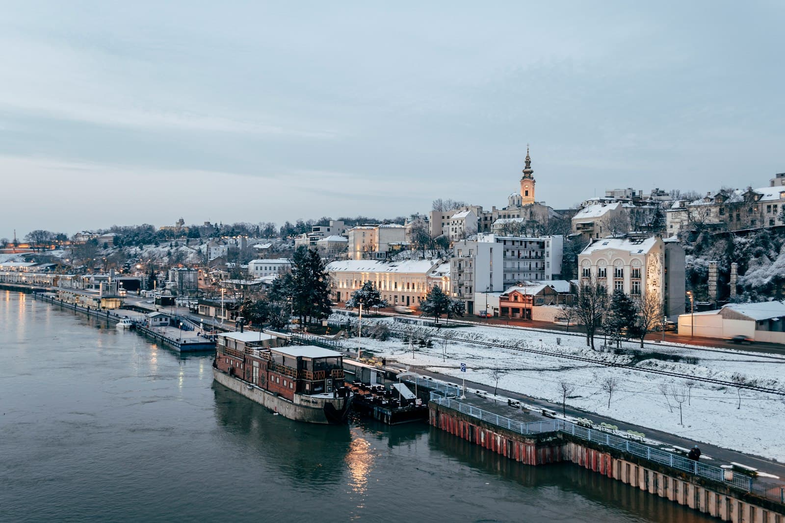 Belgrade's Sava riverfront on a snowy winter evening, with river barges and the old town above