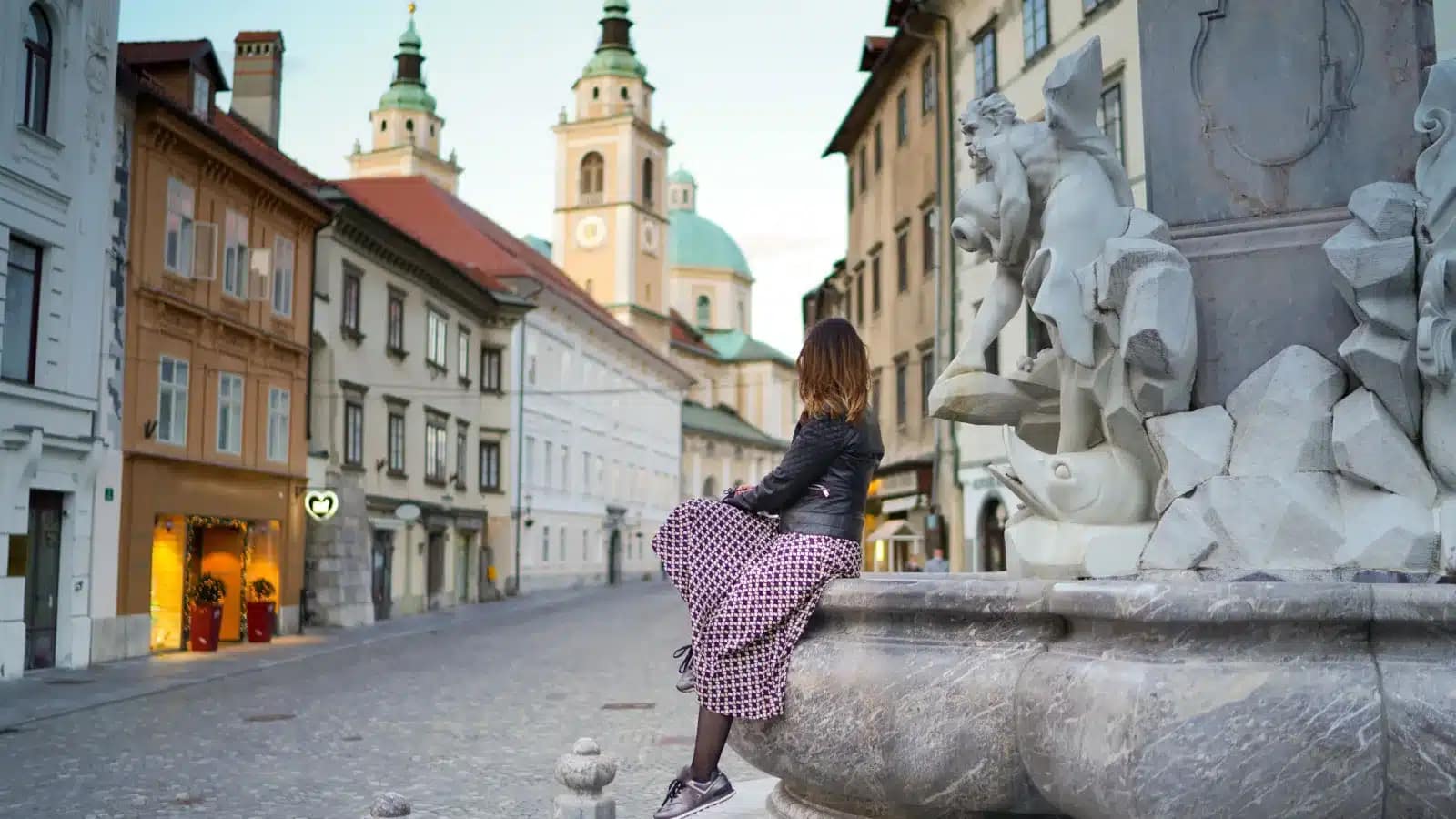 A woman sitting on the edge of the Robba Fountain in Ljubljana's old town, with the cathedral towers behind her