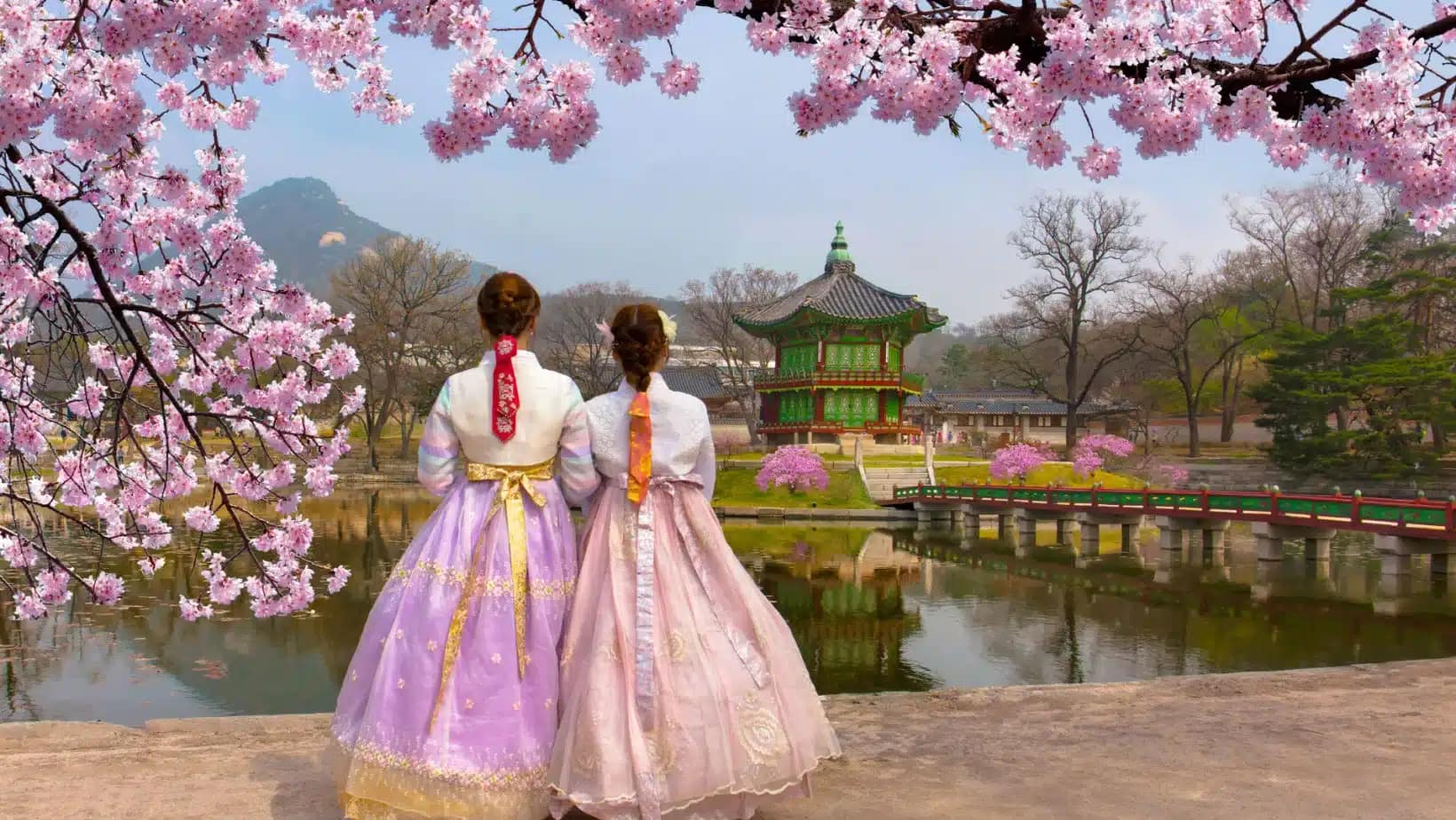 Two women in hanbok under cherry blossoms facing a pavilion on a pond at Gyeongbokgung Palace in Seoul