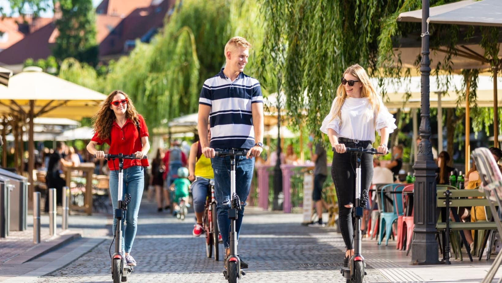 Friends riding scooters along a tree-lined riverside street of cafes in Ljubljana