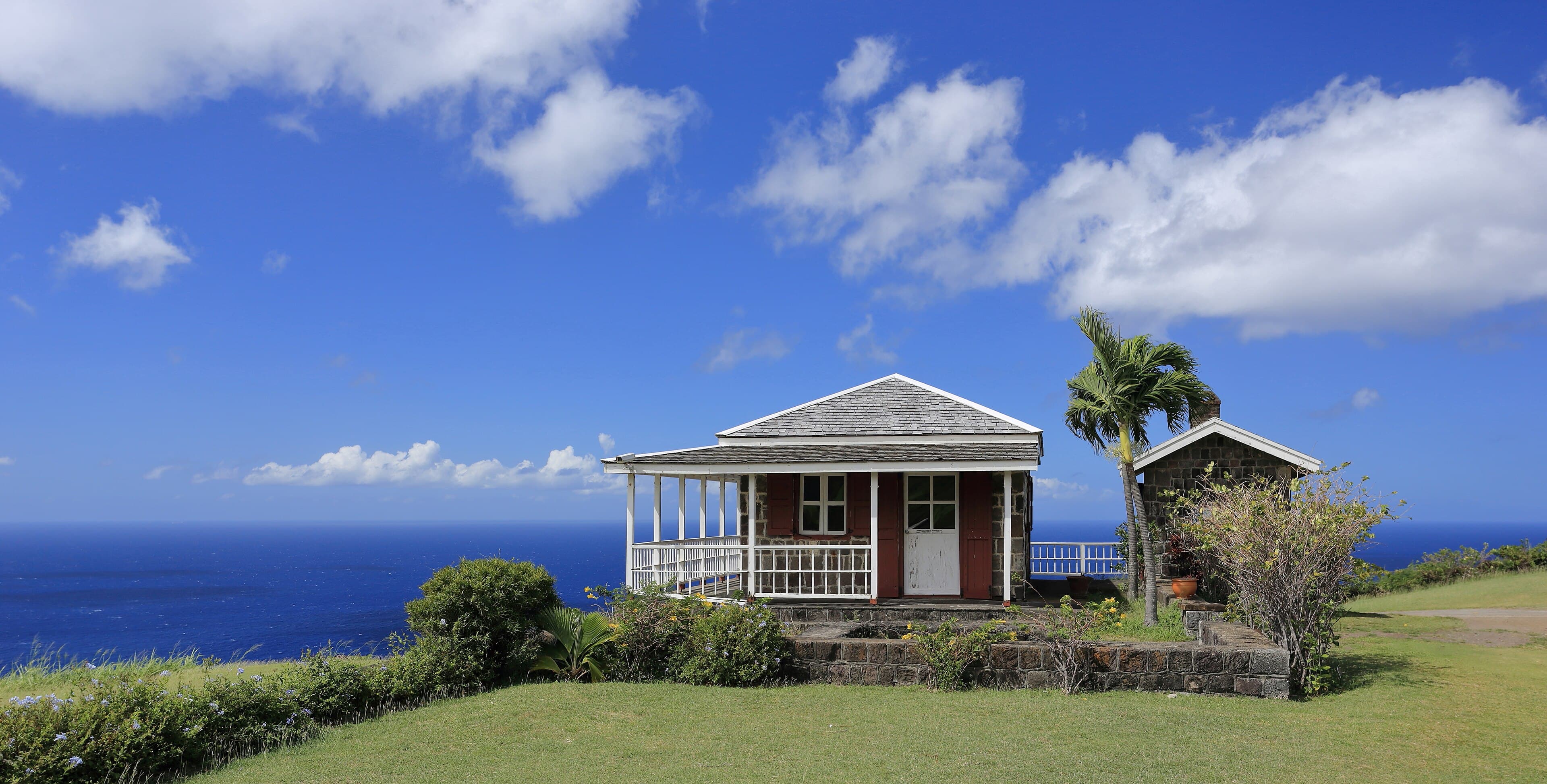 Stone cottage with a white veranda and a palm tree on a grassy hilltop above the sea at Brimstone Hill, St. Kitts