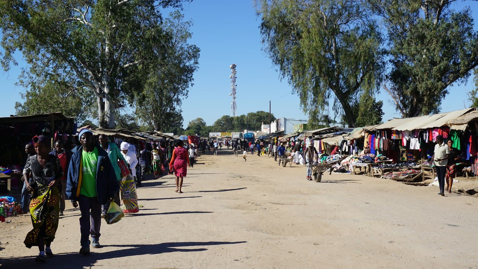 Shoppers walking down a wide dirt street lined with market stalls under a clear sky