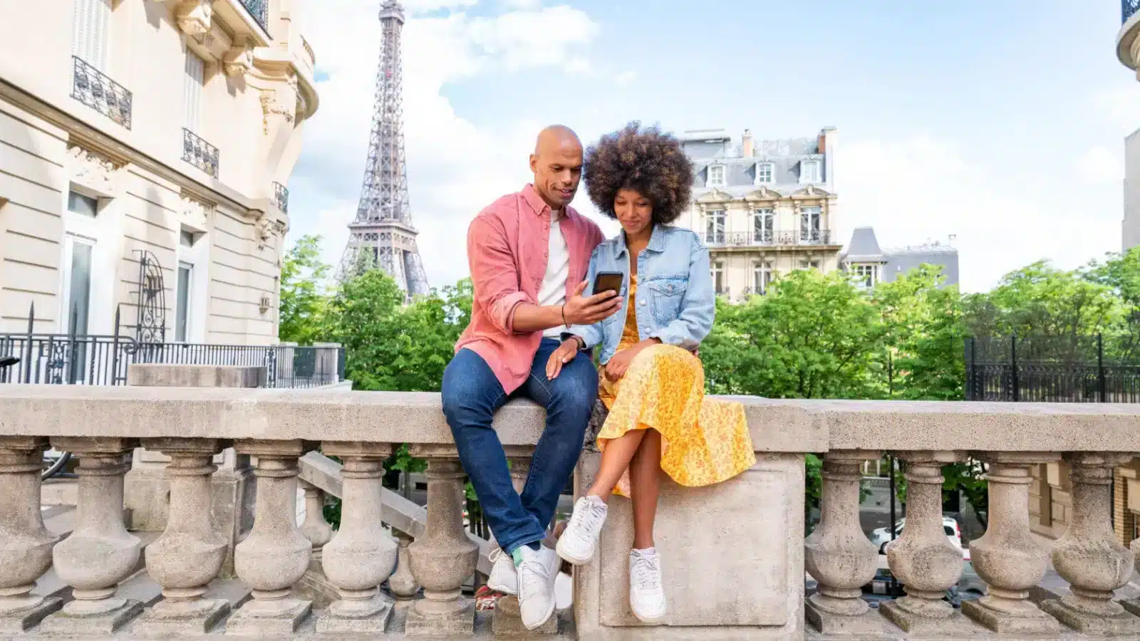 A couple sits on a bridge railing in Paris and looks at a smartphone