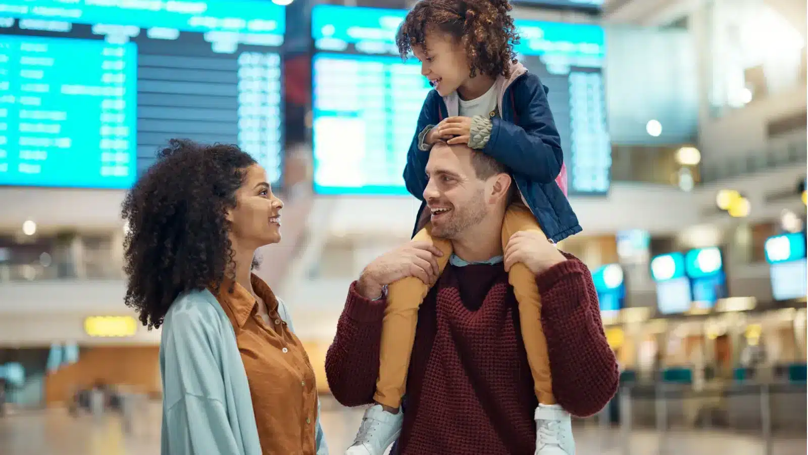 Parents at an airport with their child on his father’s shoulders, the departures board behind them